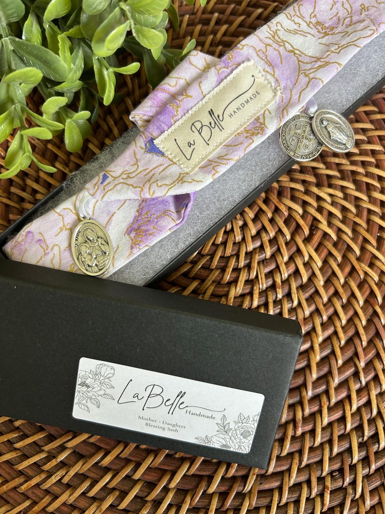Puede incluir: Una faja con estampado floral en una caja negra. La faja es de color lila claro y blanco con detalles dorados. Dos colgantes plateados cuelgan de la faja. La caja tiene una etiqueta que dice "La Belle Handmade" y "Mother + Daughter Blessing Sash."