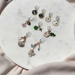 May include: A collection of nine small metal charms with clear plastic clips, featuring various designs including a black sun, a black smiley face, a white sun, a white moon, a pink flower, a green flower, a star, a shell, and a butterfly.