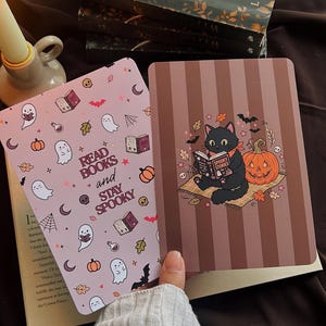 Könnte beinhalten: Zwei rechteckige Lesezeichen mit Halloween-Motiven. Eines ist hellrosa mit Geistern, Kürbissen, Fledermäusen und dem Text "READ BOOKS and STAY SPOOKY". Das andere hat braune vertikale Streifen, eine lesende schwarze Katze und einen Kürbis.