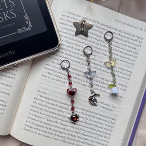 Puede incluir: Tres dijes de metal con anillas, que presentan un diseño de corazón rojo, una mariposa con una luna creciente y una mariposa con una flor blanca. Estos dijes se muestran sobre la página de un libro abierto junto a un lector Kindle.