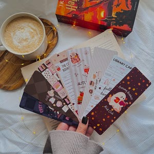 Könnte beinhalten: Eine Sammlung von sechs rechteckigen Lesezeichen aus Papier mit verschiedenen herbstlichen Illustrationen, darunter ein lesendes Gespenst, Bibliothekskartendesigns und Kaffeemotive, die in einer Hand vor einem Buch und einer Tasse aufgefächert sind.
