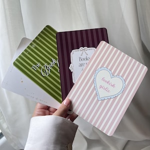 Peut inclure: Trois cartes rectangulaires aux coins arrondis. L'une est rose clair avec des rayures blanches et un cœur central portant l'inscription bookish girlie. Une autre est violet foncé rayé avec du texte. La troisième est verte rayée avec du texte.