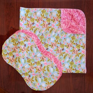 May include: A baby blanket and two matching burp cloths. The set features a light blue background with a floral pattern in shades of pink, green, and brown. The edges of the burp cloths and the blanket's trim are pink.