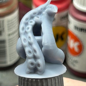 May include: A light gray, 3D-printed miniature of an octopus tentacle, featuring suckers and a curved shape. The tentacle is mounted on a circular base, with a dark gray cylindrical support. The background shows blurred paint bottles.