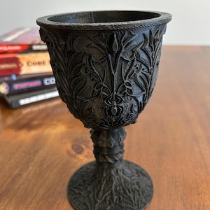 May include: A black goblet with an intricate, detailed design. The goblet has a wide, round bowl and a tall, slender stem. The base of the goblet is also decorated with a similar design.