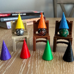 May include: A collection of colorful, miniature dunce caps, each with the word "DUNCE" printed on them, alongside various dice. The caps are in yellow, orange, blue, purple, red, green, and black. Some are placed on tiny chairs.