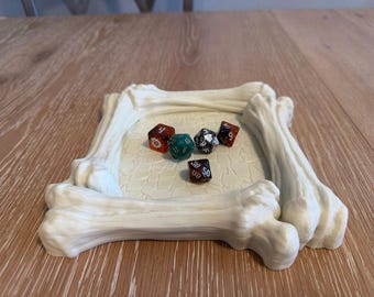 May include: A cream-colored, bone-shaped dice tray with six multi-sided dice inside. The tray is square with rounded corners, and the dice are various colors, including red, green, and black. The tray is on a wooden surface.