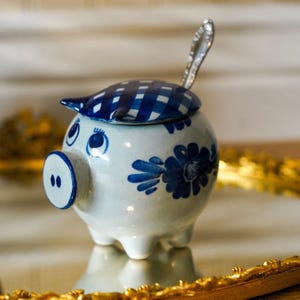 May include: A ceramic pig-shaped condiment jar with a white base and blue hand-painted floral motifs. The lid features a blue and white checkered pattern, and a silver-toned metal spoon is inserted through a small opening in the top of the lid.
