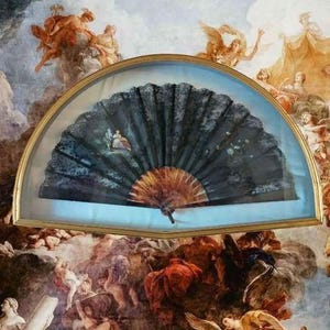 May include: A black folding hand fan with a painted scene of a seated figure, encased in a semi-circular gold-toned frame. The fan features intricate dark lace-like detailing and a light brown wooden base, displayed against a mural background.