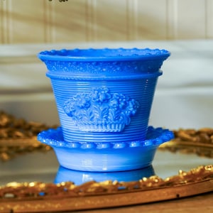 May include: A vibrant cobalt blue glass flower pot with a matching saucer. The pot features a horizontal ribbed texture and a raised relief design of a basket filled with flowers on the front. The rim and saucer edge have a beaded pattern.