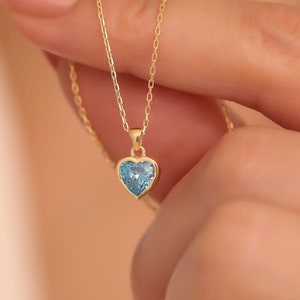May include: A delicate gold necklace featuring a heart-shaped pendant with a light blue gemstone. The pendant is suspended from a fine chain, creating a simple yet elegant design. The necklace is held up against a soft background.