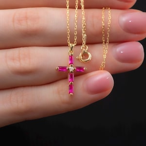 May include: A gold chain necklace with a pink gemstone cross pendant. The cross has a small clear gemstone in the centre.