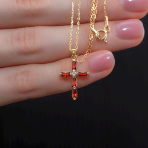 May include: A gold chain necklace with a red and white cross pendant. The cross is adorned with small red stones.