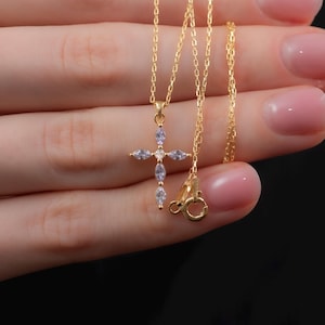 May include: A gold chain necklace with a small, clear crystal cross pendant. The necklace is held in a hand with manicured nails.