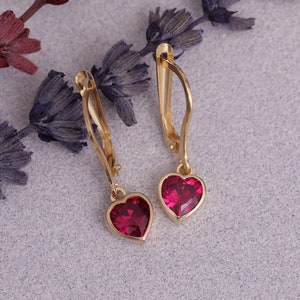 Op de afbeelding: Een paar gouden hartvormige oorbellen met rode edelsteenaccenten. De oorbellen hebben een eenvoudig, elegant ontwerp.