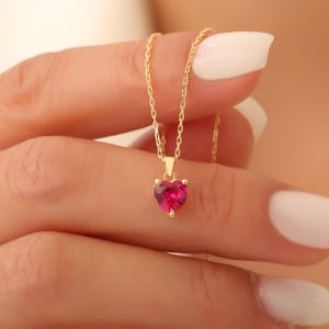 Könnte beinhalten: Eine goldene Kettenkette mit einem rosa Herz-Anhänger. Das Herz ist mit einem funkelnden rosa Stein besetzt.