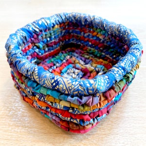 May include: A square-shaped woven basket made from colourful fabric scraps. The basket has a blue fabric rim with a gold pattern and is woven with a variety of colours, including red, orange, yellow, green, blue, and purple.