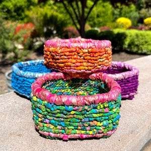 May include: Four colorful woven baskets made from fabric scraps. The baskets are stacked on top of each other. The top basket is orange and pink, the second basket is blue, the third basket is green and pink, and the bottom basket is purple.