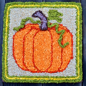 May include: A hand-stitched rug depicting a pumpkin with green leaves. The pumpkin is made of orange and red fabric and is surrounded by a green and yellow border.