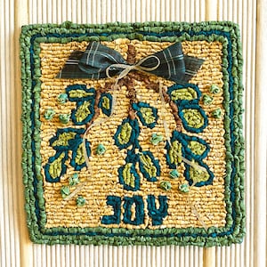 Puede incluir: Una alfombra verde y marrón con un diseño floral y la palabra "JOY" en el centro. La alfombra está hecha de tela y tiene un borde con flecos.