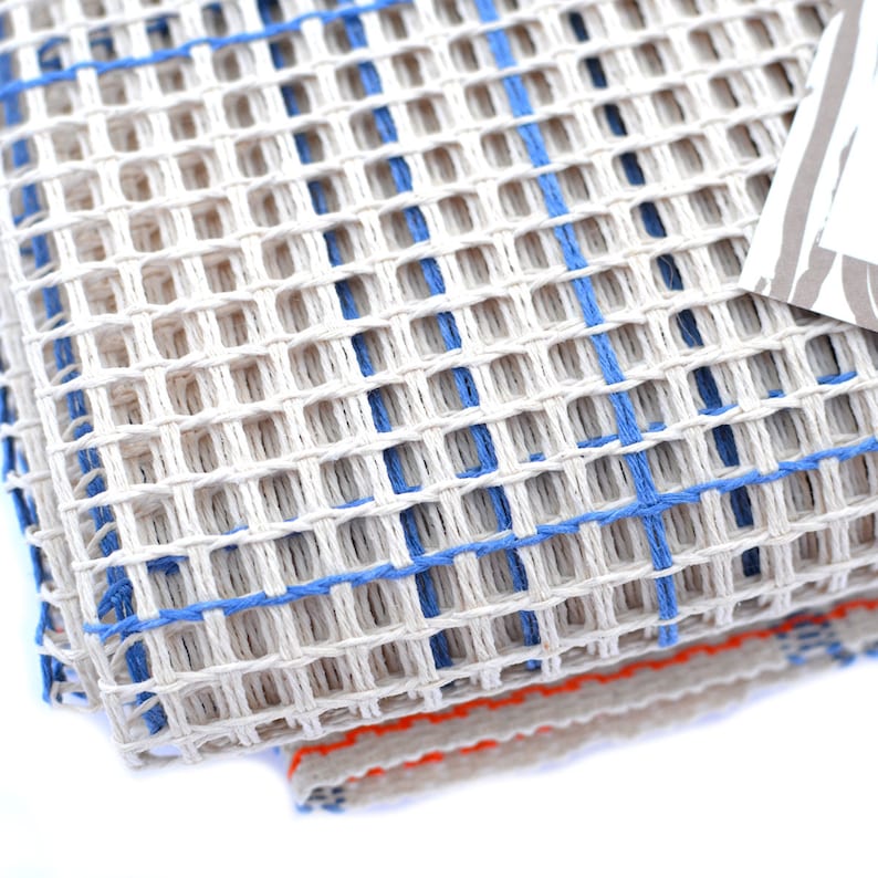 May include: A close-up of a white canvas with blue and orange threads woven into a grid pattern. The canvas is folded in half, revealing the orange threads underneath.
