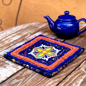 May include: A blue and orange crocheted pot holder with a colorful floral tile design in the center. The pot holder is sitting on a wooden table next to a blue teapot.