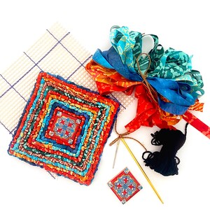 May include: A colorful rug hooking kit with a pre-made rug, yarn, ribbon, and a needle. The rug is a square with a red, orange, yellow, blue, and green pattern. The yarn is black. The ribbon is a mix of blue, green, and orange. The needle is gold.