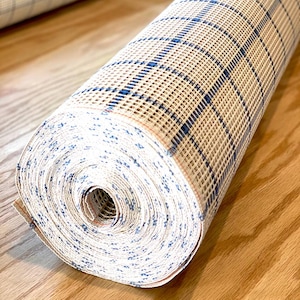 May include: Rolled rug canvas with a grid pattern in beige and blue. The canvas is rolled up, revealing the pattern and texture. The rug is on a wooden surface.
