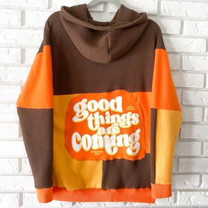 May include: A brown, orange, and yellow patchwork hoodie with a white graphic that says "good things are coming" on the back.