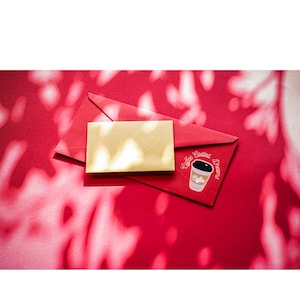 Könnte beinhalten: Ein roter Umschlag mit einer goldenen Karte und einem Aufkleber einer Kaffeetasse mit den Worten "Coffee Coated Dreams". Die Artikel befinden sich auf einer roten Oberfläche mit Schatten.