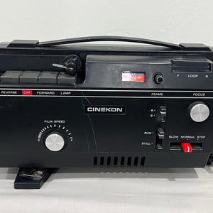 May include: A black Cinekon film projector featuring a top carrying handle, a film speed dial, and various control switches labeled Reverse, Off, Forward, Lamp, Frame, Focus, Run, Still, Slow, Normal, and Step, with a red switch and silver knobs.