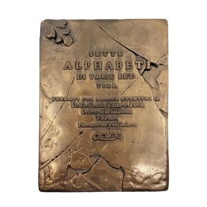 May include: A rectangular bronze-colored plaque with a cracked stone texture. It features embossed text reading SETTE ALPHABETI DI VARIE LETTERE, FORMATI CON RAGION GEOMETRIA, Da Ferdinando Ruano c. Pacen, scrittor della biblioteca Vaticana, Nuouamente posti in luce.