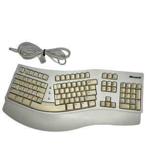 May include: A beige Microsoft ergonomic keyboard with a curved, split-key layout and an integrated wrist rest. The device features a standard QWERTY key arrangement, a numeric keypad, and a coiled white cable with a circular connector plug.
