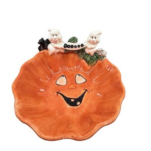 May include: A ceramic pumpkin-shaped bowl featuring a hand-painted smiling face with two teeth. The rim is decorated with two white ghosts holding a banner that reads BOOOOO, a small black cat, a grey mouse, and green leaves.