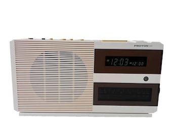 May include: A white Proton 320 clock radio featuring a horizontal slatted speaker grille on the left side, a digital display showing 12:03, and a brown-tinted radio tuner window below the display with various frequency markings printed on it.