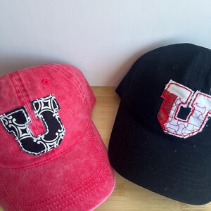 May include: A red baseball cap and a black baseball cap, each featuring a large, fabric-stitched letter U on the front. The red cap has a black and white patterned letter, while the black cap has a white and red patterned letter.