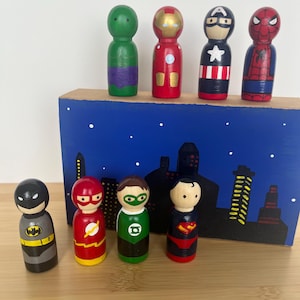 May include: A set of eight wooden superhero figurines, painted in various colours, including red, blue, green, yellow, and black. The figurines are standing on a wooden base with a painted cityscape backdrop.