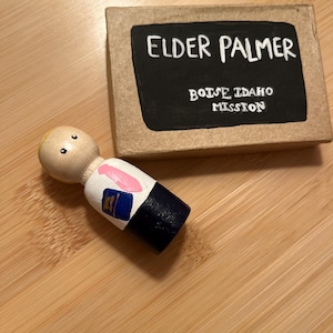 May include: A wooden figurine of a missionary wearing a white shirt, a pink tie, and a blue suit jacket. The figurine is holding a book. A small sign reads "Elder Palmer Boise Idaho Mission".