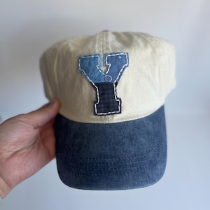 May include: A baseball cap with a cream-colored crown and a dark blue, distressed-look brim. The front features a large, appliquéd letter Y constructed from multiple blue fabric patches with visible white hand-stitched detailing around the edges.