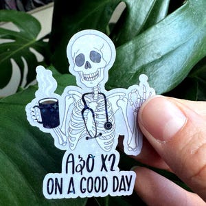 May include: A sticker featuring a skeleton with a stethoscope, holding a mug of coffee. The sticker has the text "A&O X2 ON A GOOD DAY". The skeleton is white with black outlines, and the coffee mug is dark blue with white stars.