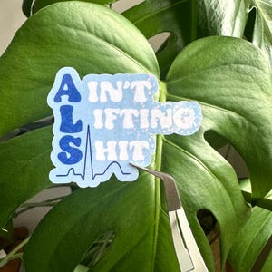 Op de afbeelding: Een blauwe sticker met de tekst "AIN'T LIFTING SHIT" in een speels lettertype, met een hartslaglijn grafiek. De sticker wordt vastgehouden door een pincet voor een groot groen blad.