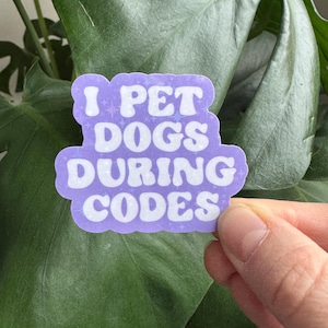 Può includere: Un adesivo viola con il testo bianco "I PET DOGS DURING CODES". L'adesivo è mostrato su uno sfondo di una grande foglia verde.