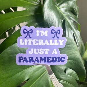 Op de afbeelding: Een sticker met de tekst "I'M LITERALLY JUST A PARAMEDIC" in wit en marineblauw, met strikaccenten. De sticker wordt vastgehouden door een pincet tegen een achtergrond van grote, groene monstera bladeren.