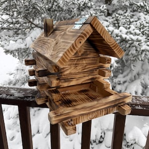 May include: A wooden bird feeder shaped like a small house with a slanted roof. The feeder is made of natural wood with a rustic finish and has a small platform for birds to perch on. The feeder is mounted on a wooden railing.
