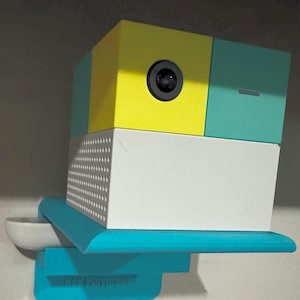 May include: A multi-colored camera with a yellow, teal, and white block design. The camera has a black lens and is mounted on a teal shelf with a small bowl. The text "Not Playground" is visible on the shelf.