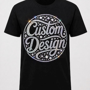 Op de afbeelding: Zwart t-shirt met een cirkelvormig ontwerp. Het ontwerp toont de woorden "Custom Design" in een cursief lettertype, omlijnd met sprankelende strass-steentjes. De strass-steentjes zijn in een cirkelvormig patroon gerangschikt, wat een glinsterend effect creëert.
