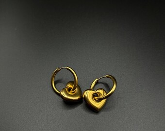 Puede incluir: Un par de pendientes de aro dorados con un dije en forma de corazón. Los aros son simples y los corazones están ligeramente elevados.