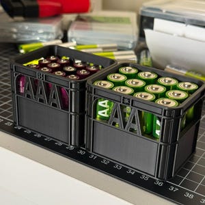 May include: Two black plastic crates filled with AAA batteries. The crates have the letters "AAA" printed on the front. One crate is filled with purple batteries and the other with green batteries.