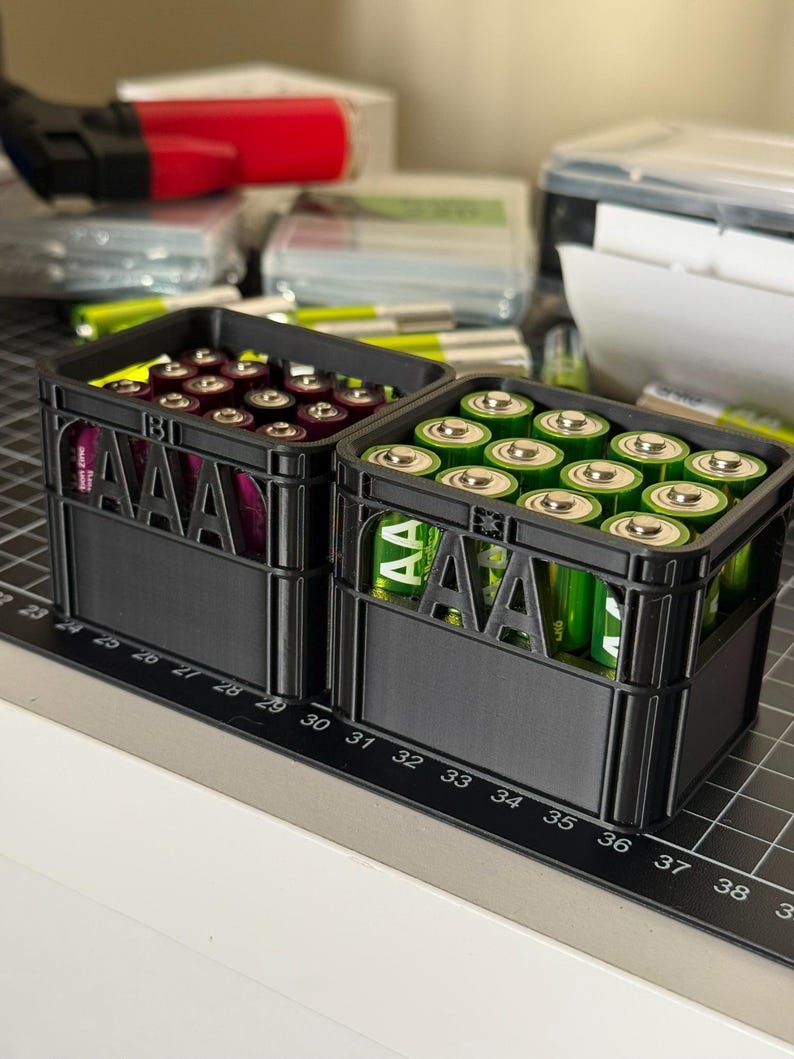 May include: Two black plastic crates filled with AAA batteries. The crates have the letters "AAA" printed on the front. One crate is filled with purple batteries and the other with green batteries.