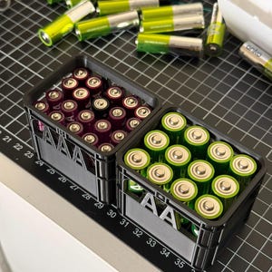 May include: Two black plastic storage containers with the label "AA" on the front. The containers are filled with batteries, one with purple AA batteries and the other with green AA batteries.